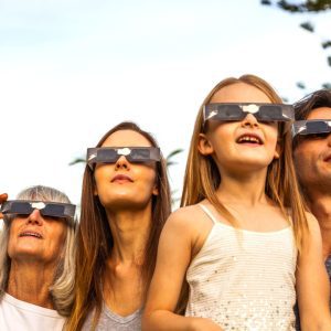 A mother, father, daughter, and grandmother look up to the sky while wearing solar eclipse glasses.