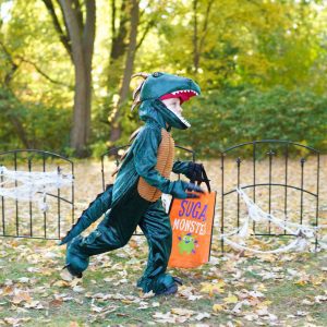 A little kid dressed as a dinosaur runs through the grounds with a trick or treat bag.