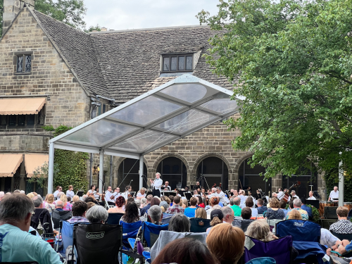 The back of Ford House with a large clear span tent. Underneath are members of the Michigan Philharmonic. In the foreground, hundreds of guests sit in camp chairs to enjoy the concert.