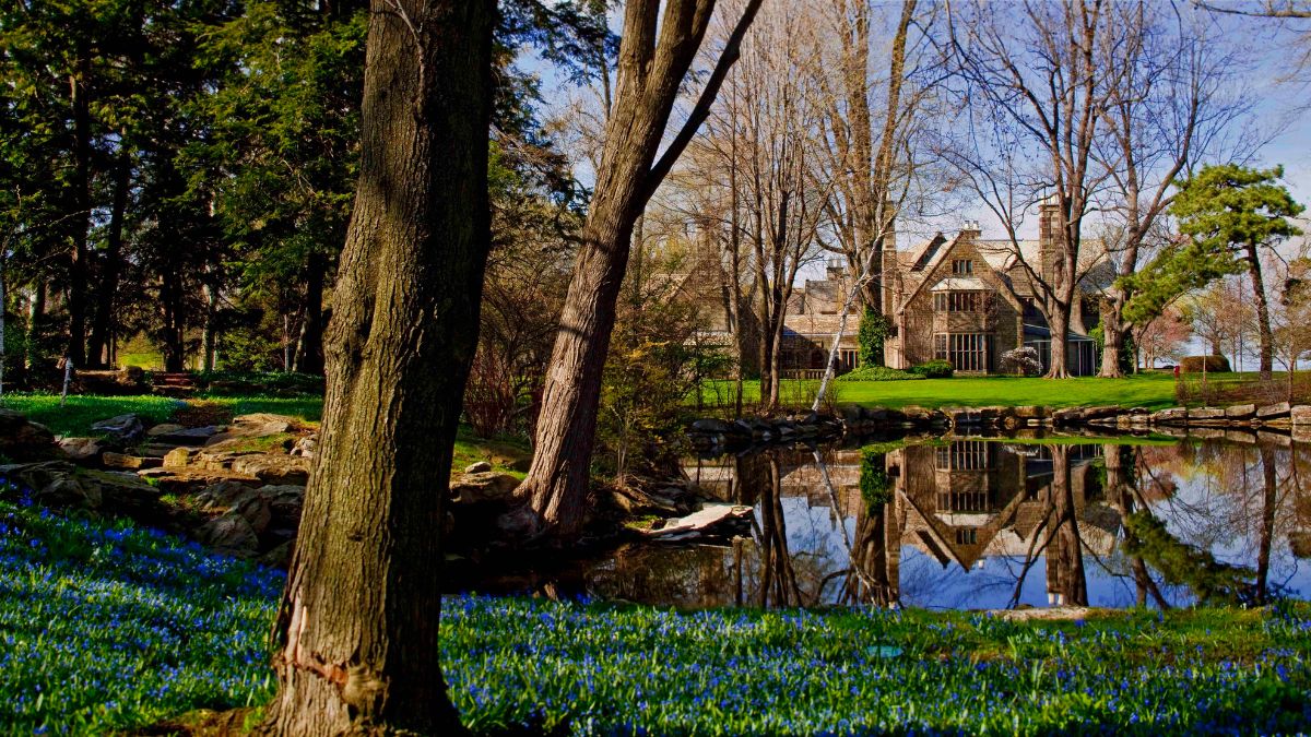 The Ford House Main Residence reflected in the calm waters of the lagoon, surrounded by early spring blooms and wooded tranquility.