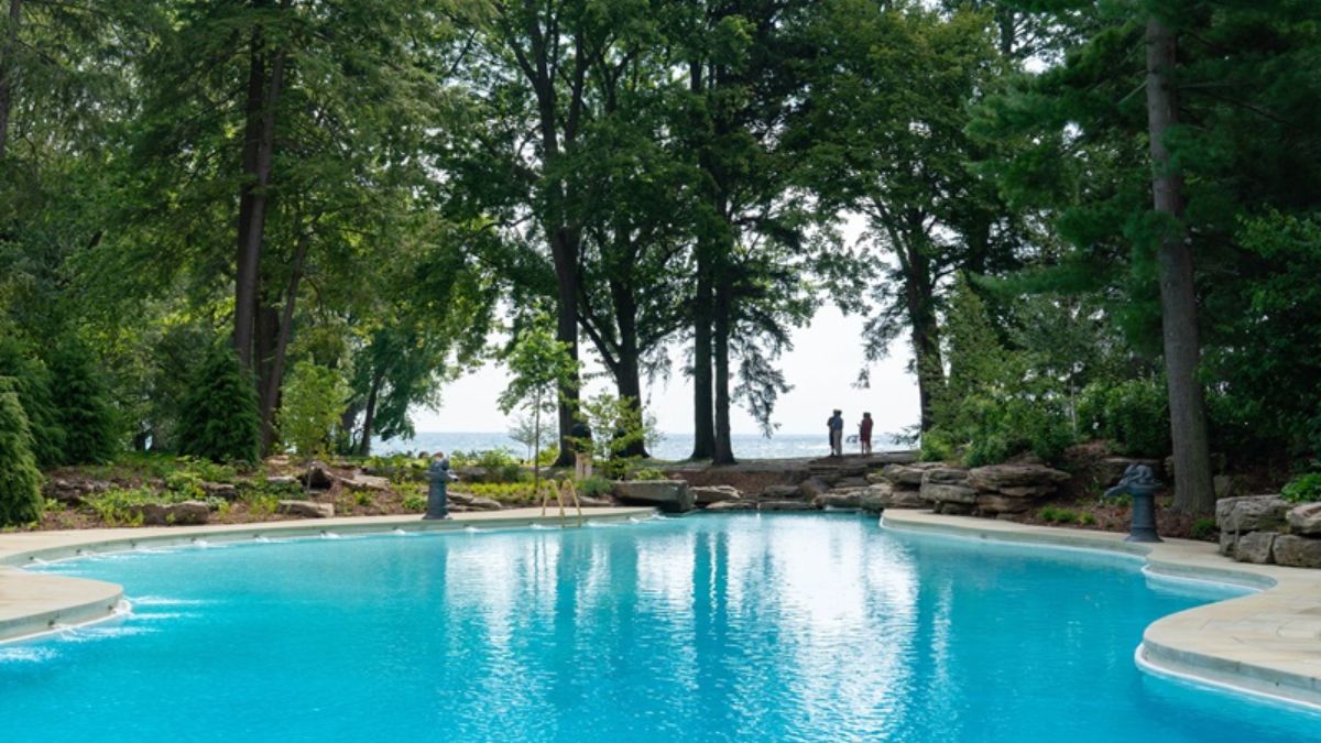 Expansive view of the Ford House pool leading toward Lake St. Clair, with tall trees and guests visible along the waterfront edge.