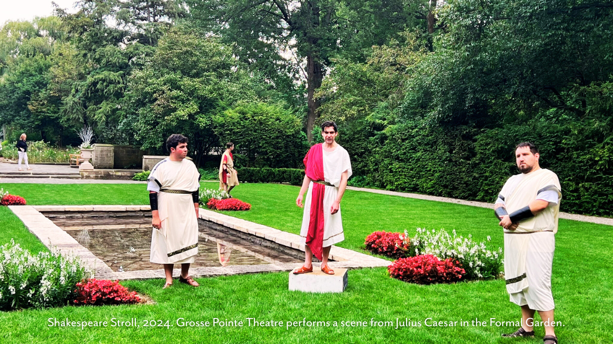 Actors from Grosse Pointe Theatre perform a scene from Julius Caesar during the 2024 Shakespeare Stroll at Ford House, standing beside the Formal Garden’s reflecting pool surrounded by vibrant flower beds and dense greenery.
