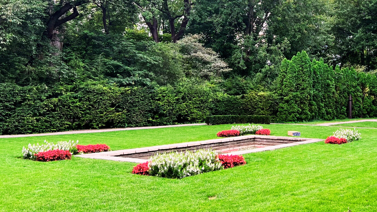 View of the Formal Garden at Ford House with a rectangular reflecting pool bordered by bright red and white flower beds, neatly trimmed grass, and tall evergreen hedges.
