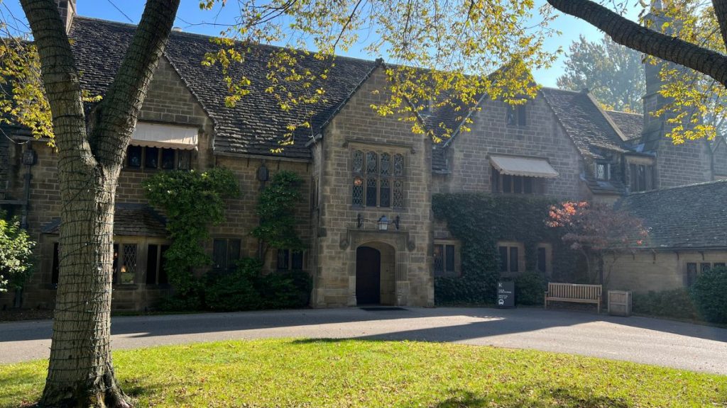Exterior of the historic Main Residence at Ford House in Grosse Pointe Shores, Michigan, featuring Tudor Revival architecture with ivy-covered stone walls, leaded glass windows, and a central arched entryway framed by mature trees and landscaped greenery on a sunny autumn day.