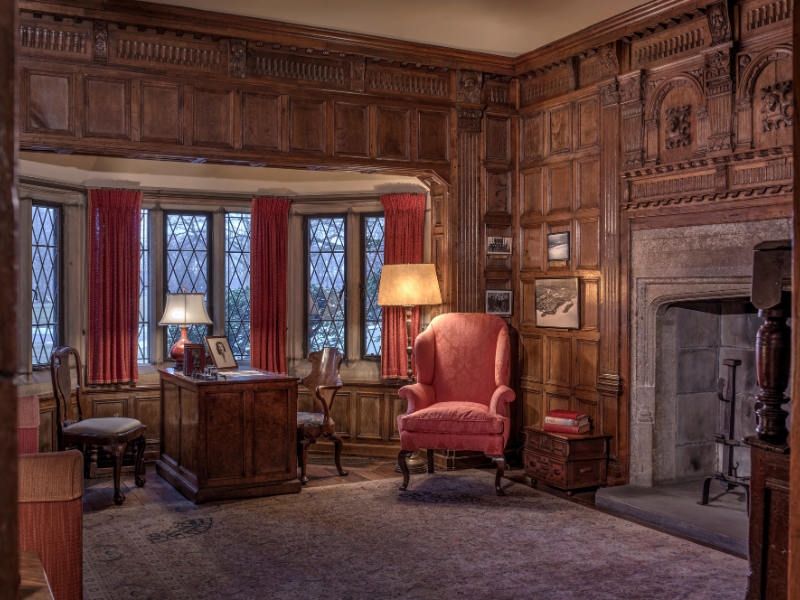 Edsel's Study. A warmly lit, wood-paneled study featuring a large bay window with diamond-patterned leaded glass panes and red drapes. In the foreground is a red wingback armchair beside a small wooden side table and floor lamp. A stone fireplace with carved wood paneling above it sits to the right, while a wooden desk with classic chairs and a table lamp occupies the center. Framed photos and books accent the space, giving it a cozy, historic ambiance.