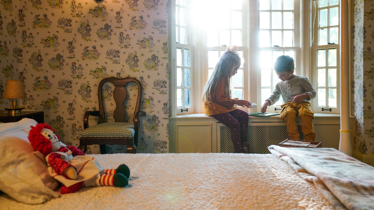 Two children read near a window seat inside Josephine’s Playhouse, surrounded by vintage wallpaper and a cozy bed with a Raggedy Ann doll.