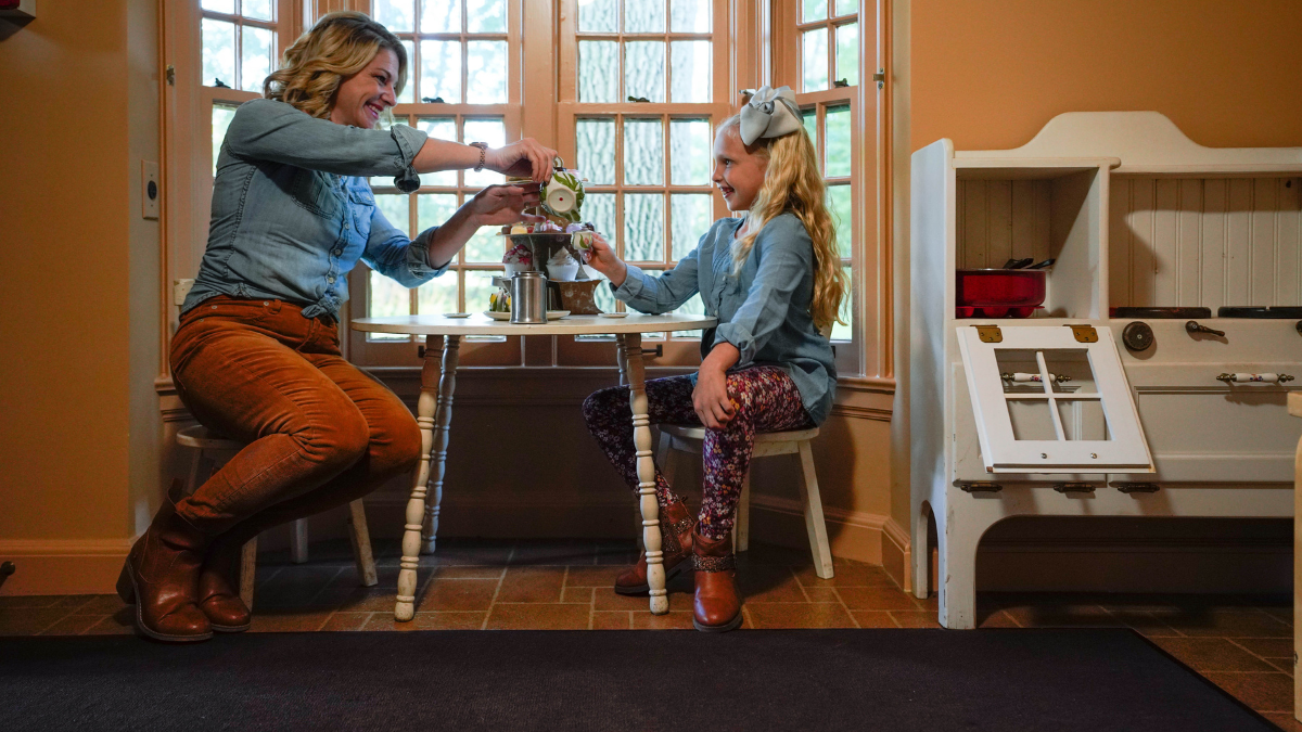 A mother and daughter share a pretend tea party at a child-sized table in Josephine Ford’s historic Playhouse, surrounded by vintage furnishings.
