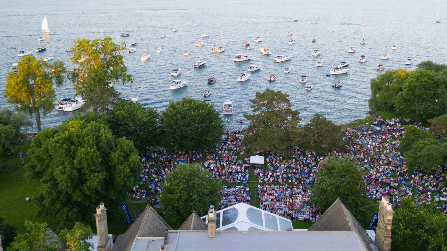 Aerial image of a massive lakeside crowd at Ford House with dozens of boats anchored on the water, watching a concert from the shoreline and from the lake.