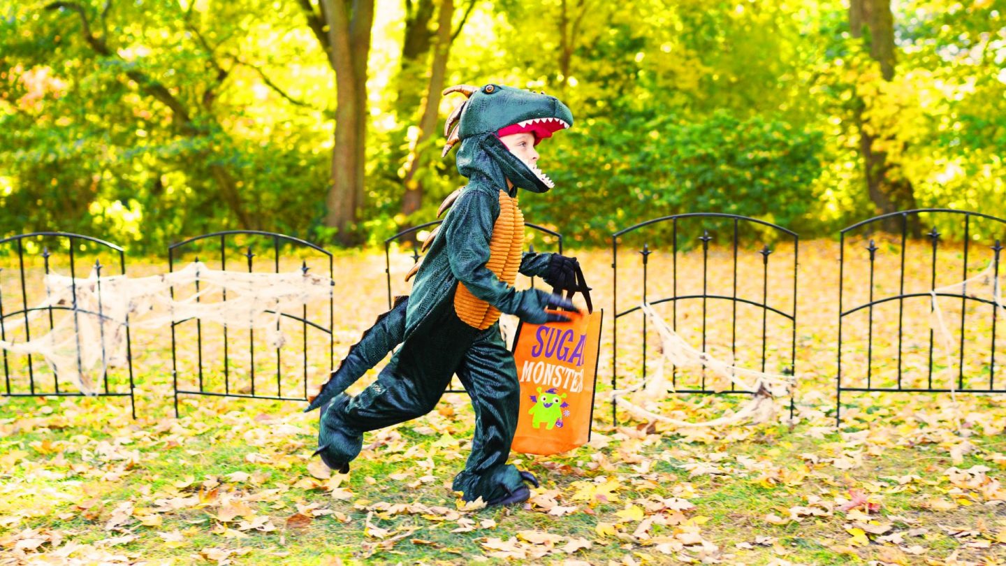 A young child in a dinosaur costume runs along a leaf-covered path with a bright orange "Sugar Monster" treat bag.