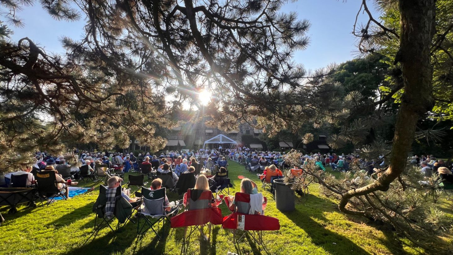 View of concertgoers seated on lawn chairs and blankets at Ford House, framed by tree branches, enjoying a live musical performance as the sun sets.