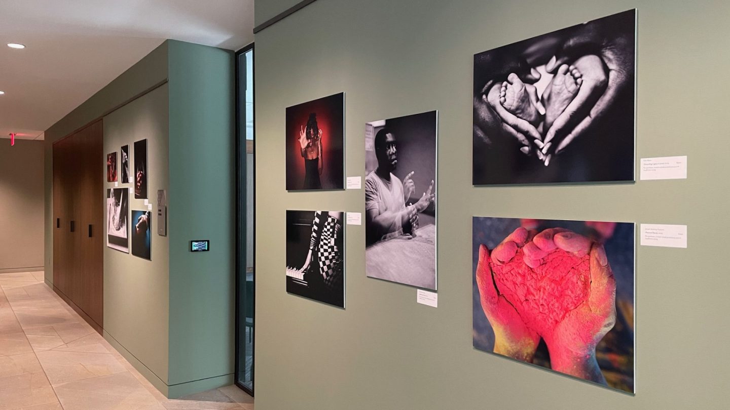 Photographs on display at Ford House's Love in Reach exhibit, including black-and-white and color images of hands forming a heart shape, vibrant red powder cupped in palms, and expressive portraits.