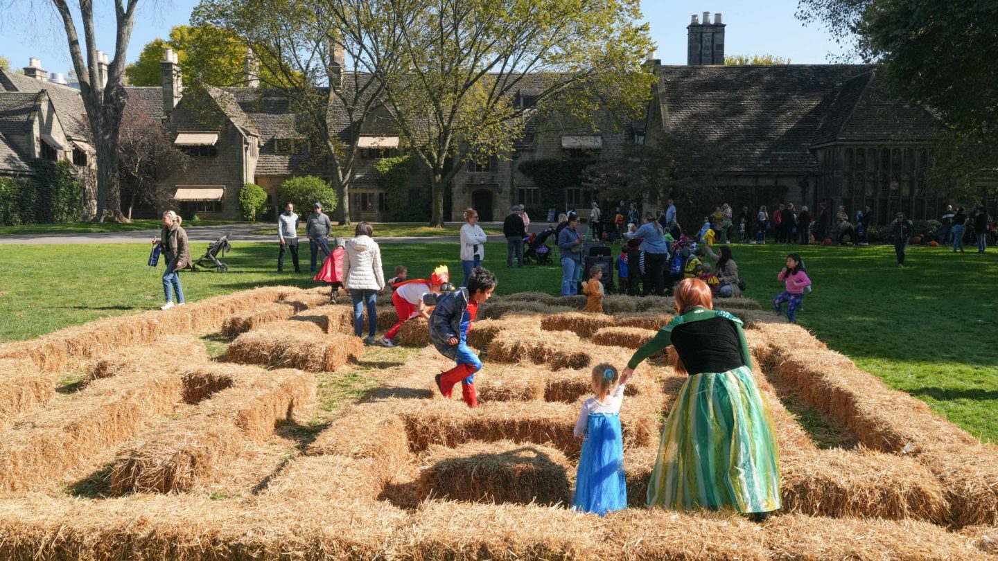 Children in costumes play in a straw bale maze on the lawn at Ford House, with the historic estate in the background.