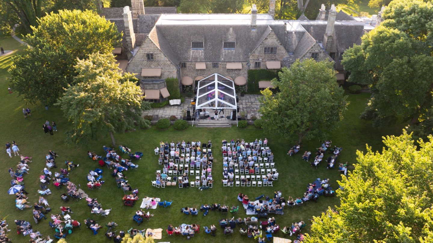 Aerial shot of a concert in progress at Ford House, with a stage tent set up on the terrace and symmetrical rows of chairs and audience members extending out onto the lawn.