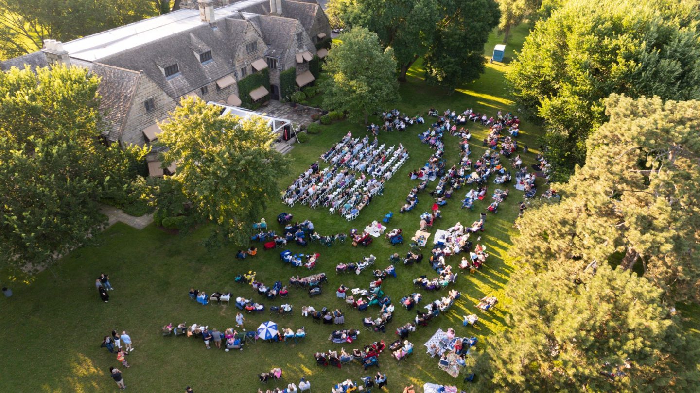 Aerial view of a large crowd gathered on the lawn at Ford House for a summer concert, with rows of white chairs and colorful picnic blankets arranged in front of the historic estate.