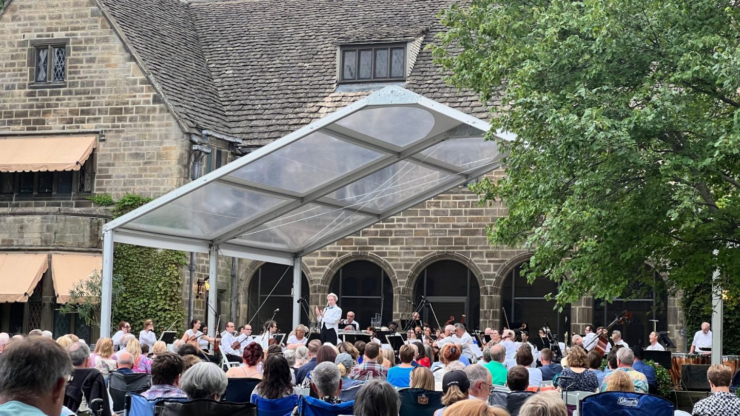 Close-up of a conductor leading an orchestra under a clear tent on the terrace of Ford House, with an attentive audience seated on the lawn.