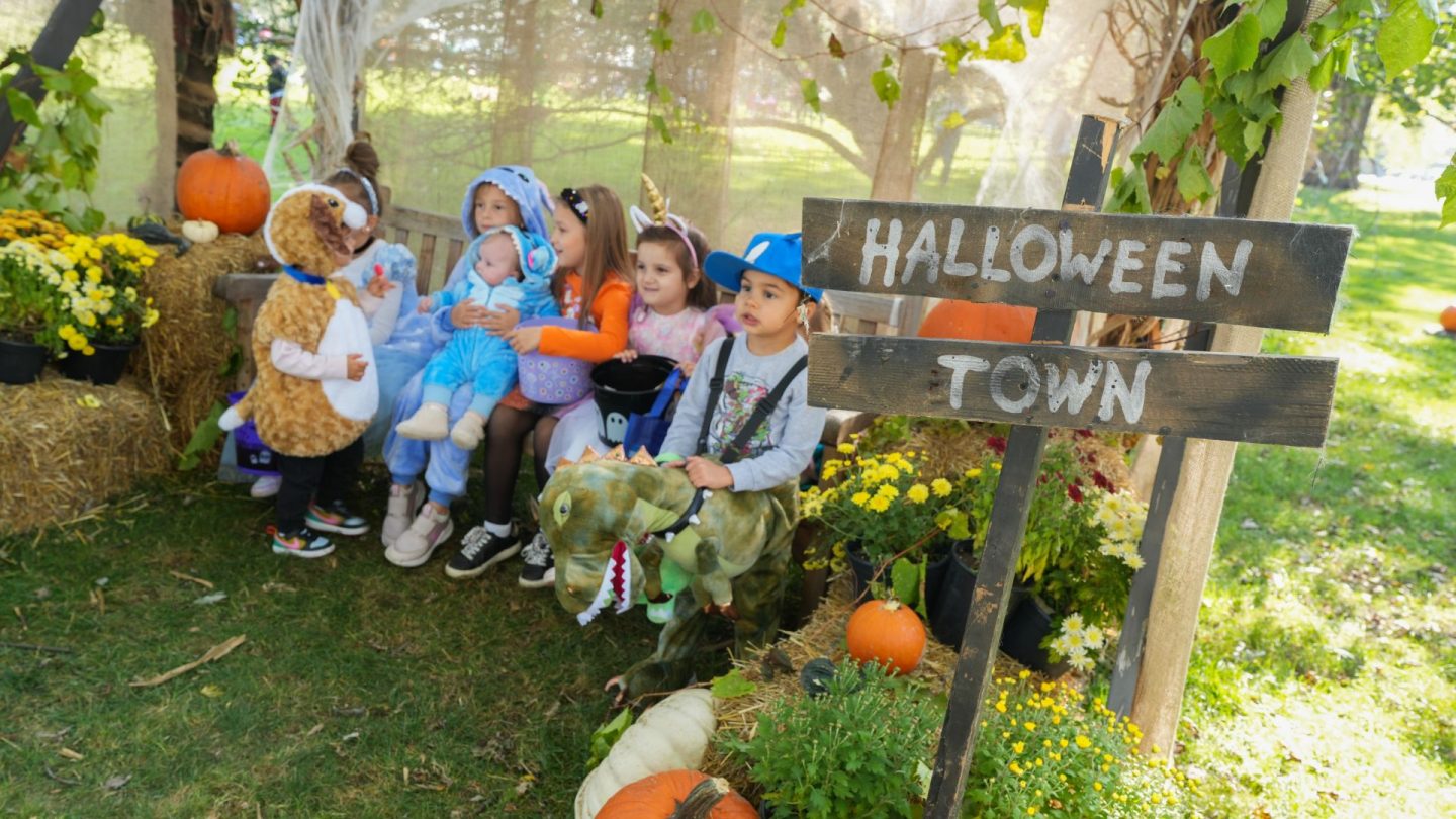A group of young children in costume gather for a photo in a cozy Halloween-themed setting marked “Halloween Town.”
