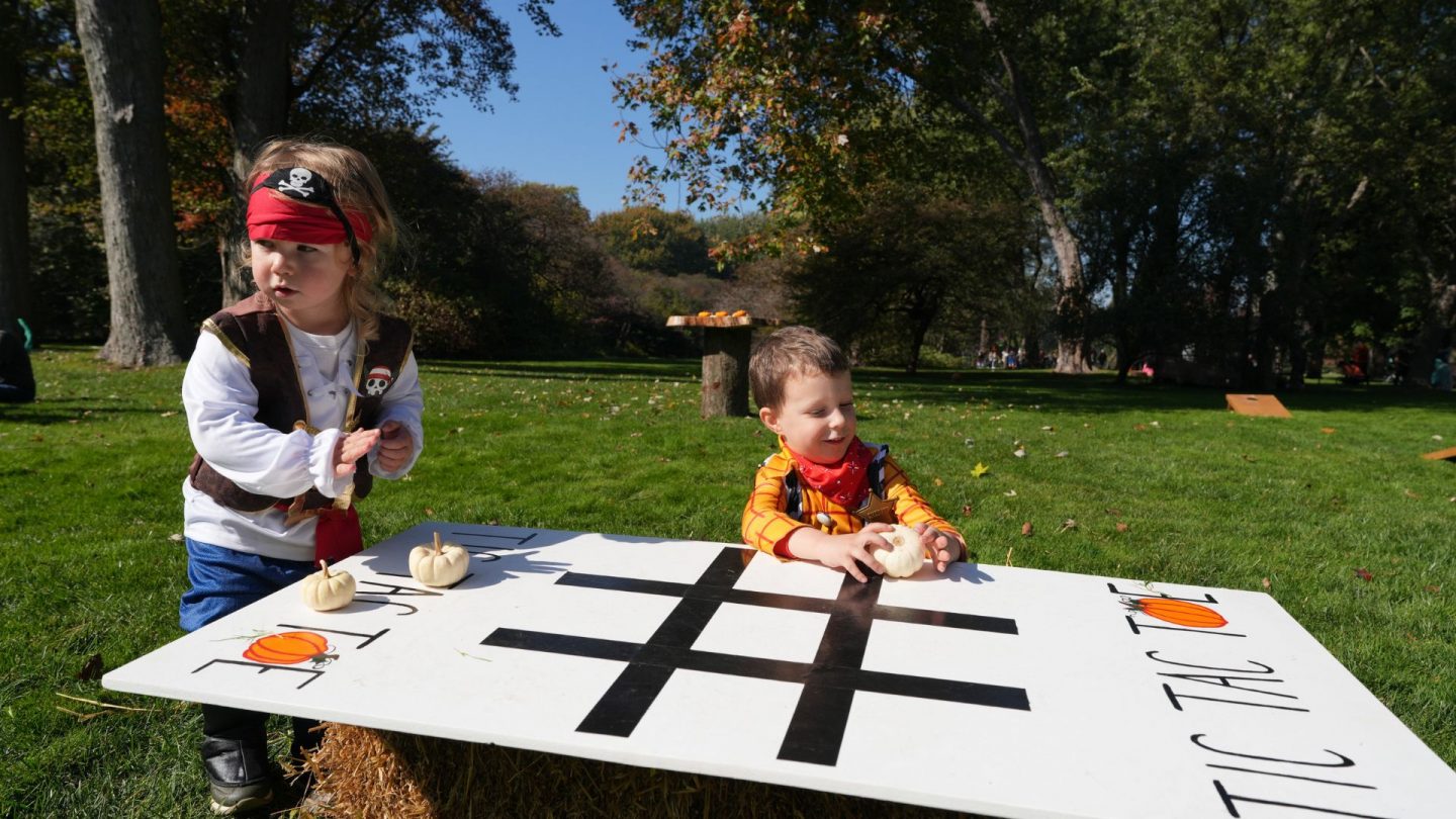 Two young children in pirate and cowboy costumes play pumpkin tic-tac-toe on the grass.