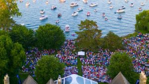 Aerial view of a large outdoor concert at Ford House with a colorful crowd seated on blankets and lawn chairs on the grass, overlooking dozens of boats anchored in the water behind the stage area.