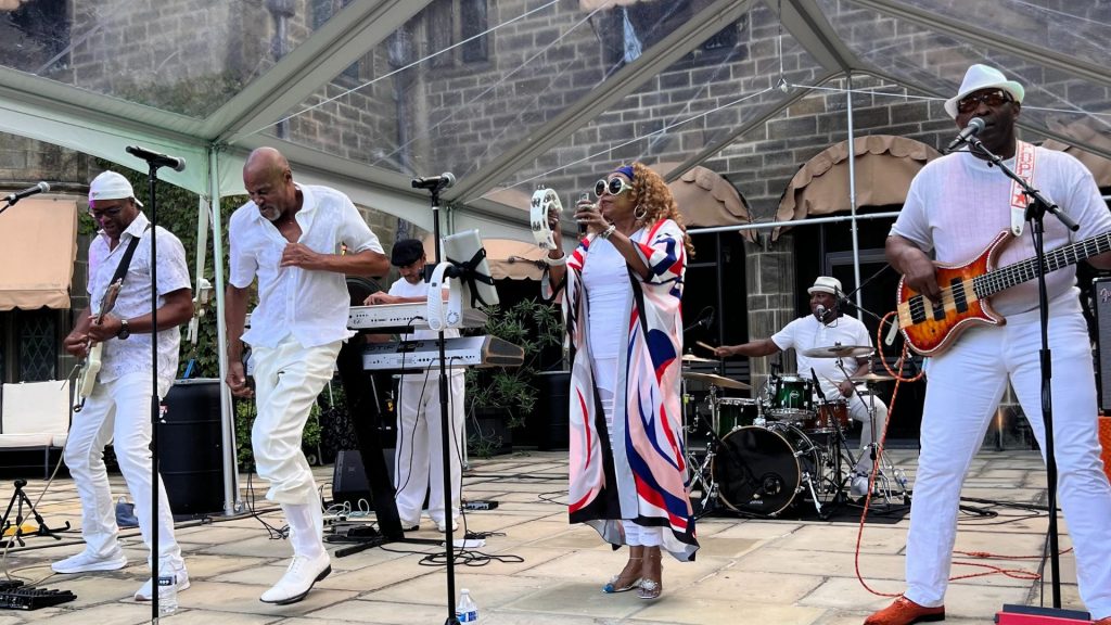 The Dave Hamilton Band performs live under a clear canopy in front of a historic stone building at Ford House. Band members, dressed in stylish white outfits, play guitar, drums, bass, keyboard, and tambourine while singing and dancing.