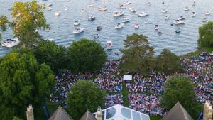Aerial view of a large crowd gathered on the lakeside lawn at Ford House during a summer concert. Dozens of boats float in Lake St. Clair beyond the shoreline as attendees sit on blankets and chairs surrounded by lush green trees.