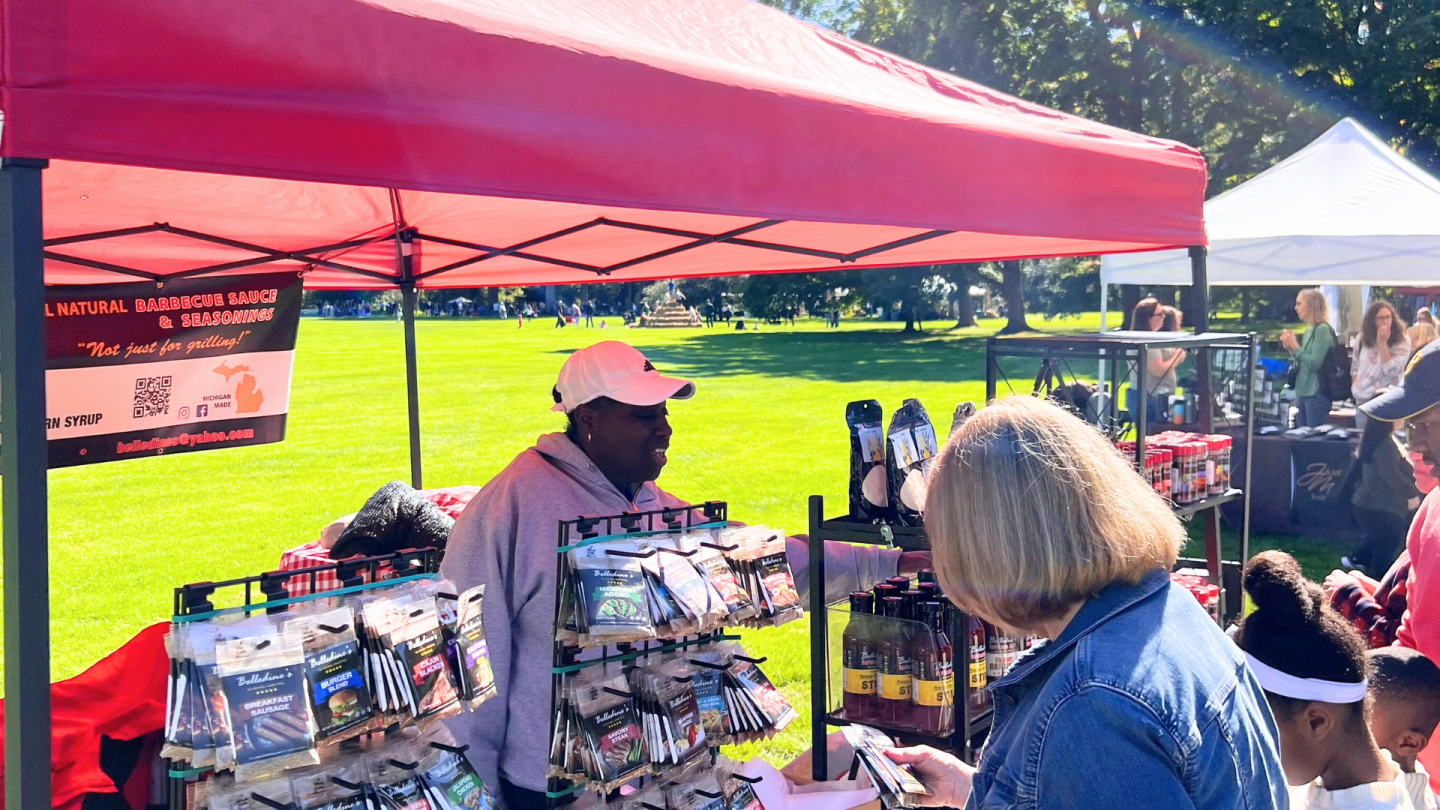 A vendor under a red canopy sells barbecue sauces and seasonings to customers at an outdoor event. Children and adults browse the booth in the sunshine.