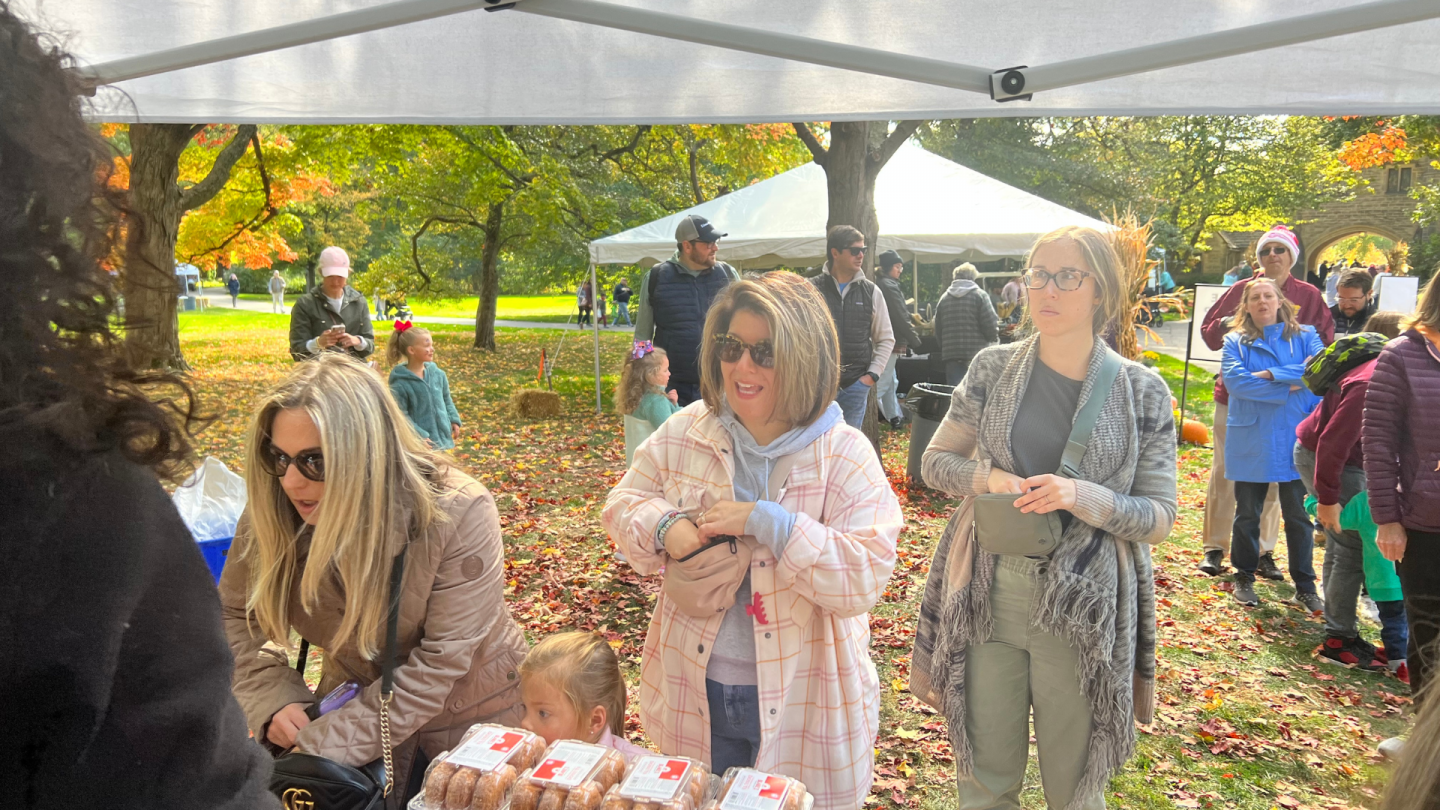 People standing under a vendor tent at an outdoor fall market; trays of cider mill donuts are visible on the table in front. Colorful autumn leaves blanket the ground around them.
