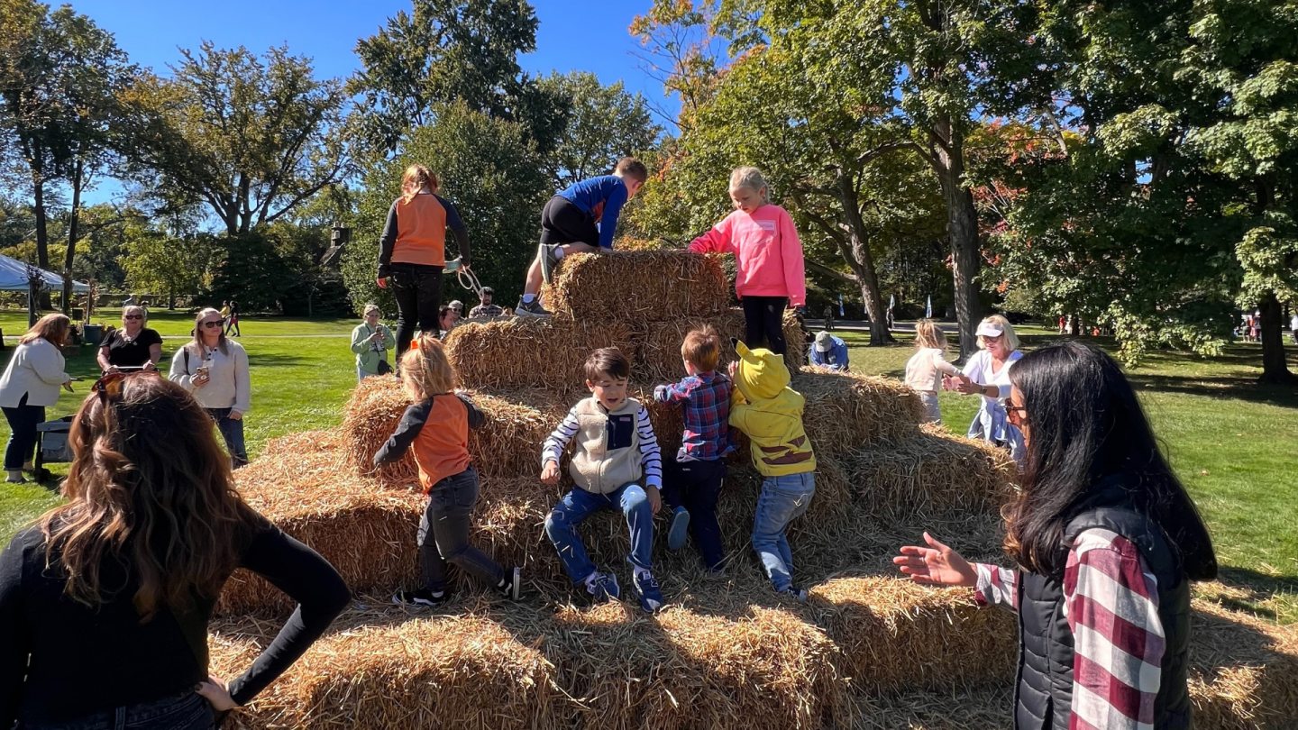 Children of various ages climb and play on a pyramid made of straw bales under the autumn sun.