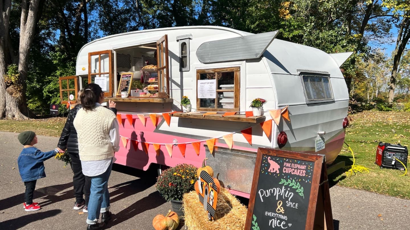 A retro-style camper turned food truck is decorated with orange bunting and a fall-themed menu board that reads “Pumpkin Spice & Everything Nice.”