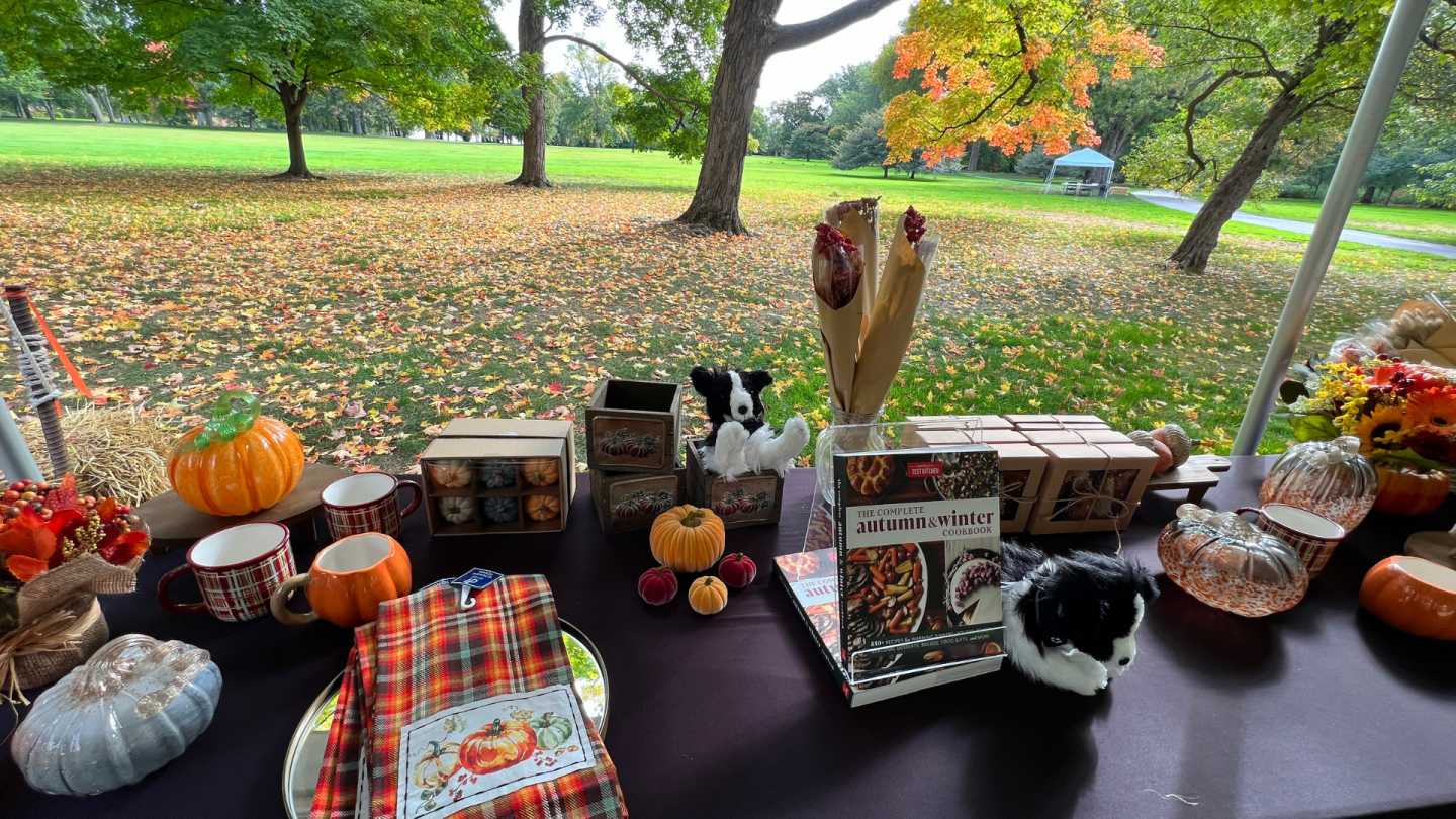 A festive vendor table displays autumn-themed items such as plaid mugs, glass pumpkins, tea towels, candles, and a cookbook. The background shows fallen leaves on a quiet grassy field.