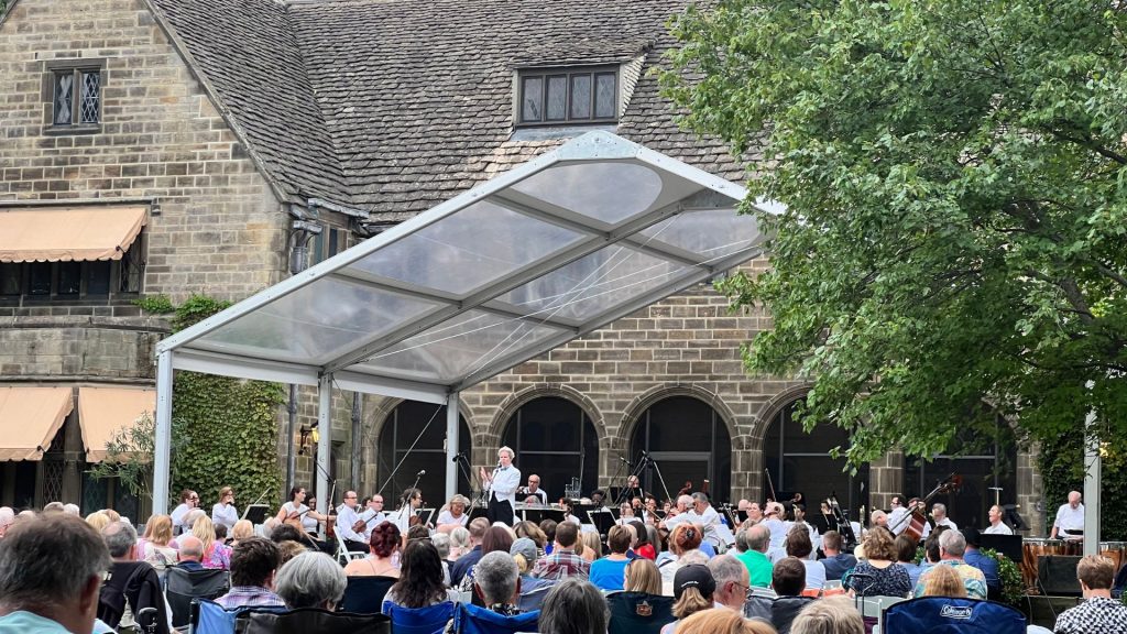 The Michigan Philharmonic orchestra performs on the terrace of Ford House beneath a large clear canopy. Musicians in black and white attire play string and percussion instruments as a conductor leads them, with a seated audience watching in front of the historic residence.