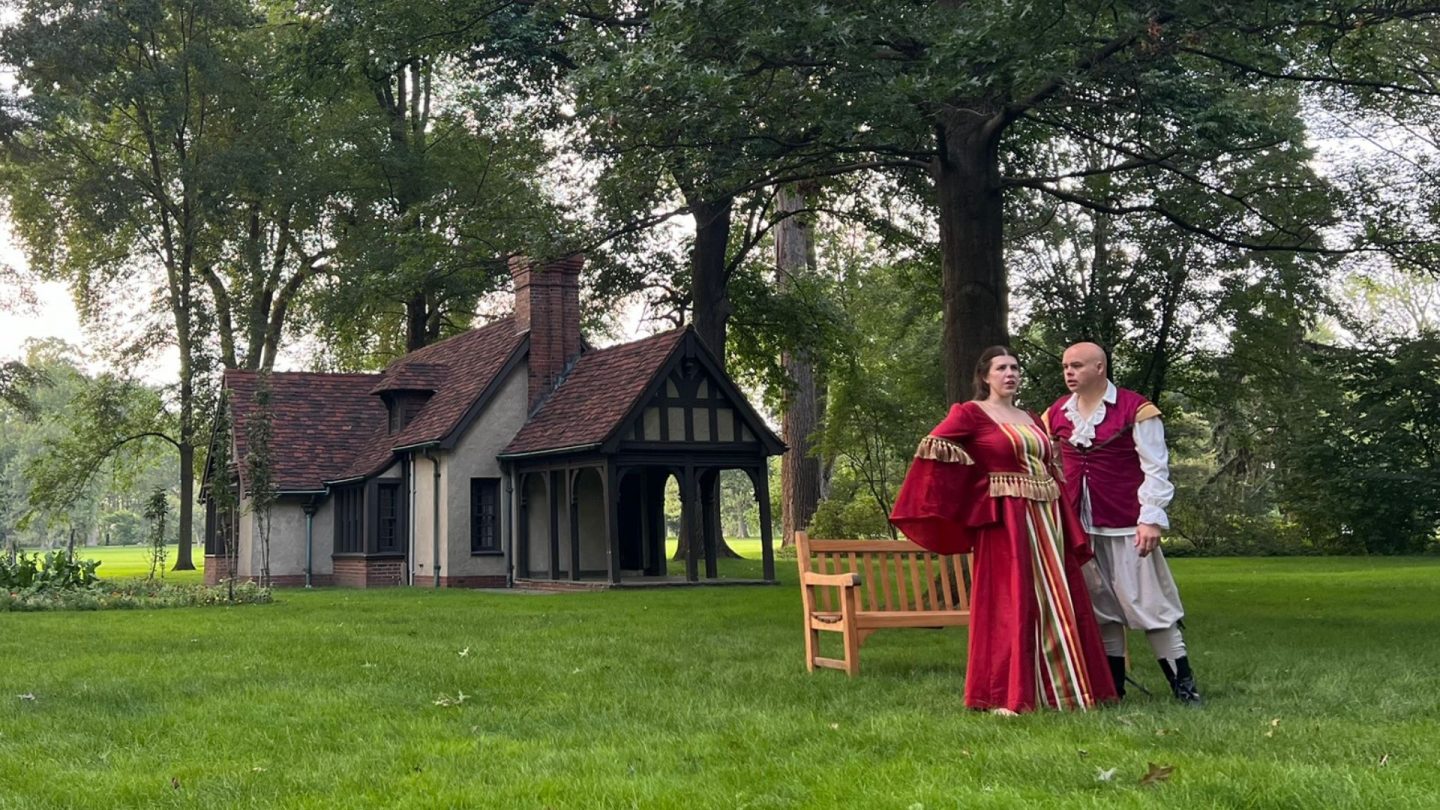 Two actors in period Renaissance costumes perform a passionate Shakespearean scene on the lawn near the Playhouse at Ford House. The woman gestures dramatically while the man looks on with intensity, backed by towering trees and the quaint historic cottage.