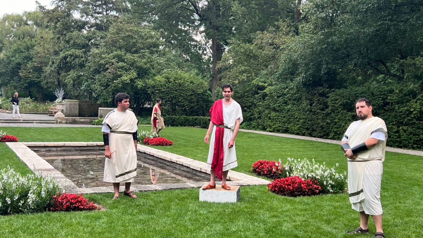 Three actors in Roman tunics deliver a scene from Julius Caesar beside the reflecting pool in Ford House’s formal garden. Red and white flowerbeds line the scene as onlookers begin to gather beyond the hedges.