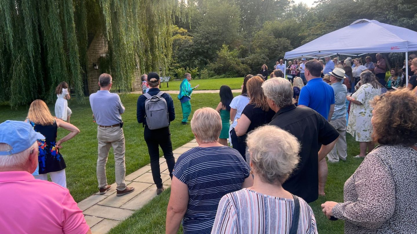 A performer in vibrant teal costume delivers a comedic Shakespearean scene under sweeping willow branches as an audience gathers on the lawn. A white tent provides shade nearby, and guests of all ages watch attentively at Ford House.