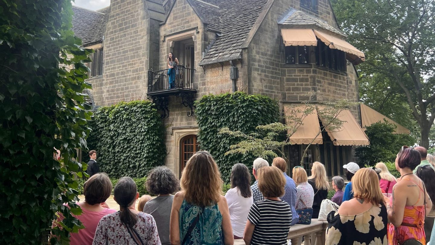 A large crowd watches an actress in blue portray Juliet from the stone balcony of the historic Ford House courtyard. Below, a male actor dressed as Romeo delivers his lines, surrounded by ivy-covered walls and period architecture.