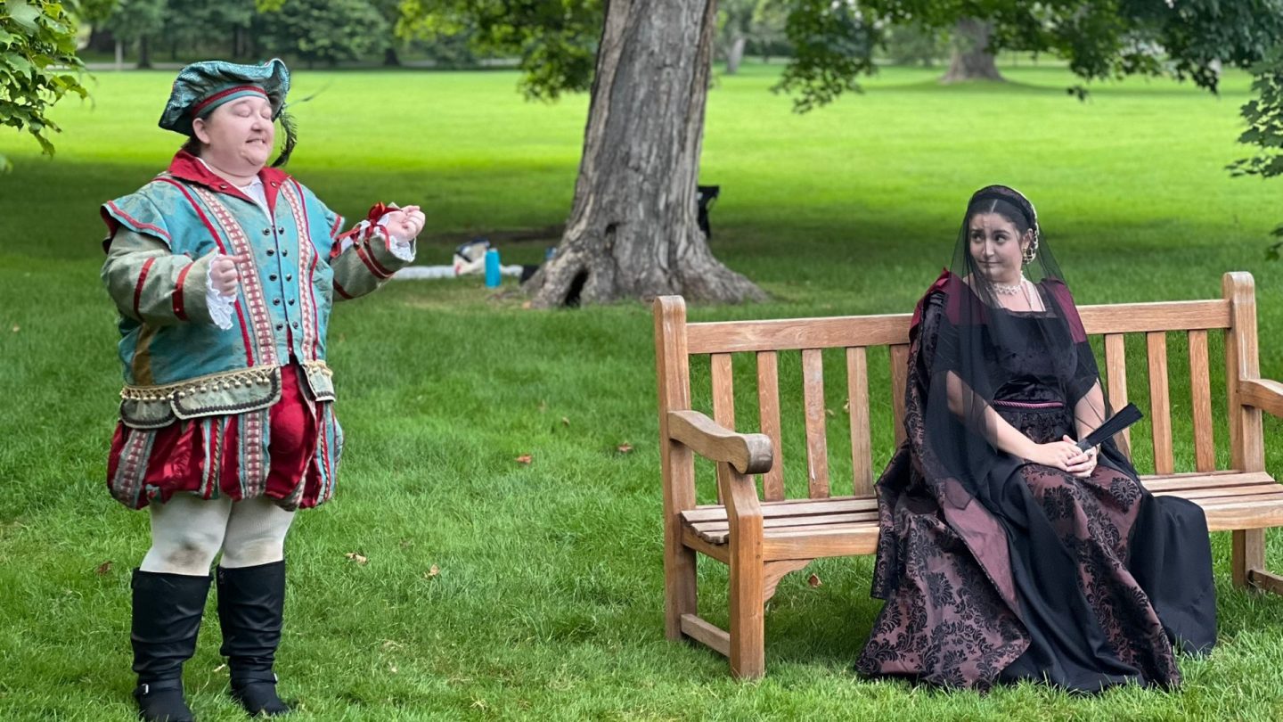 A court jester in a richly detailed teal and red costume performs with animated gestures before a seated noblewoman dressed in black and burgundy, complete with pearls and a mourning veil, during a Shakespearean scene on the Ford House lawn.