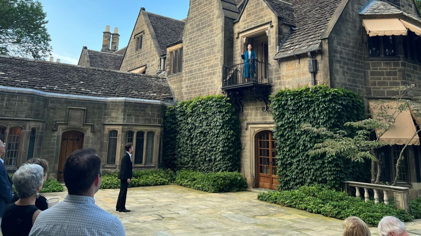 An actress in a teal gown performs Juliet’s balcony monologue from above as guests gather in the historic stone courtyard of Ford House. A male actor looks up from below in the iconic Romeo and Juliet scene, surrounded by lush ivy and elegant architecture.