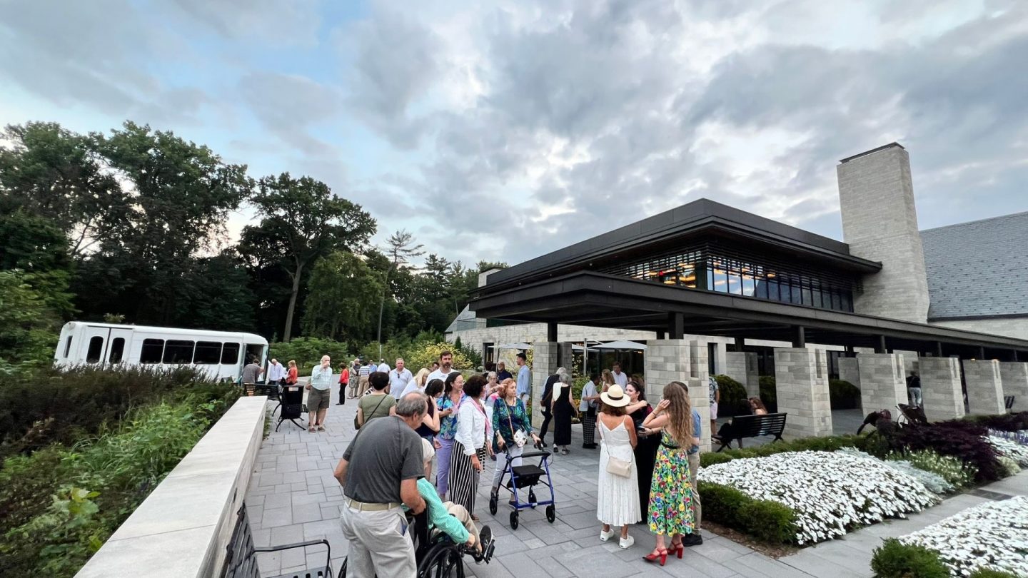 Guests arrive and mingle outside the modern Visitor Center at Ford House before the Shakespeare Stroll begins. A white shuttle bus and staff greet attendees under a dramatic sky, with flower beds and trees framing the entry walkway.