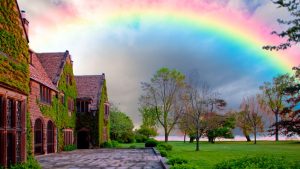 A vibrant rainbow stretches across the sky above the historic Ford House, a Tudor-style mansion covered in ivy with a stone walkway leading to the lawn and Lake St. Clair.